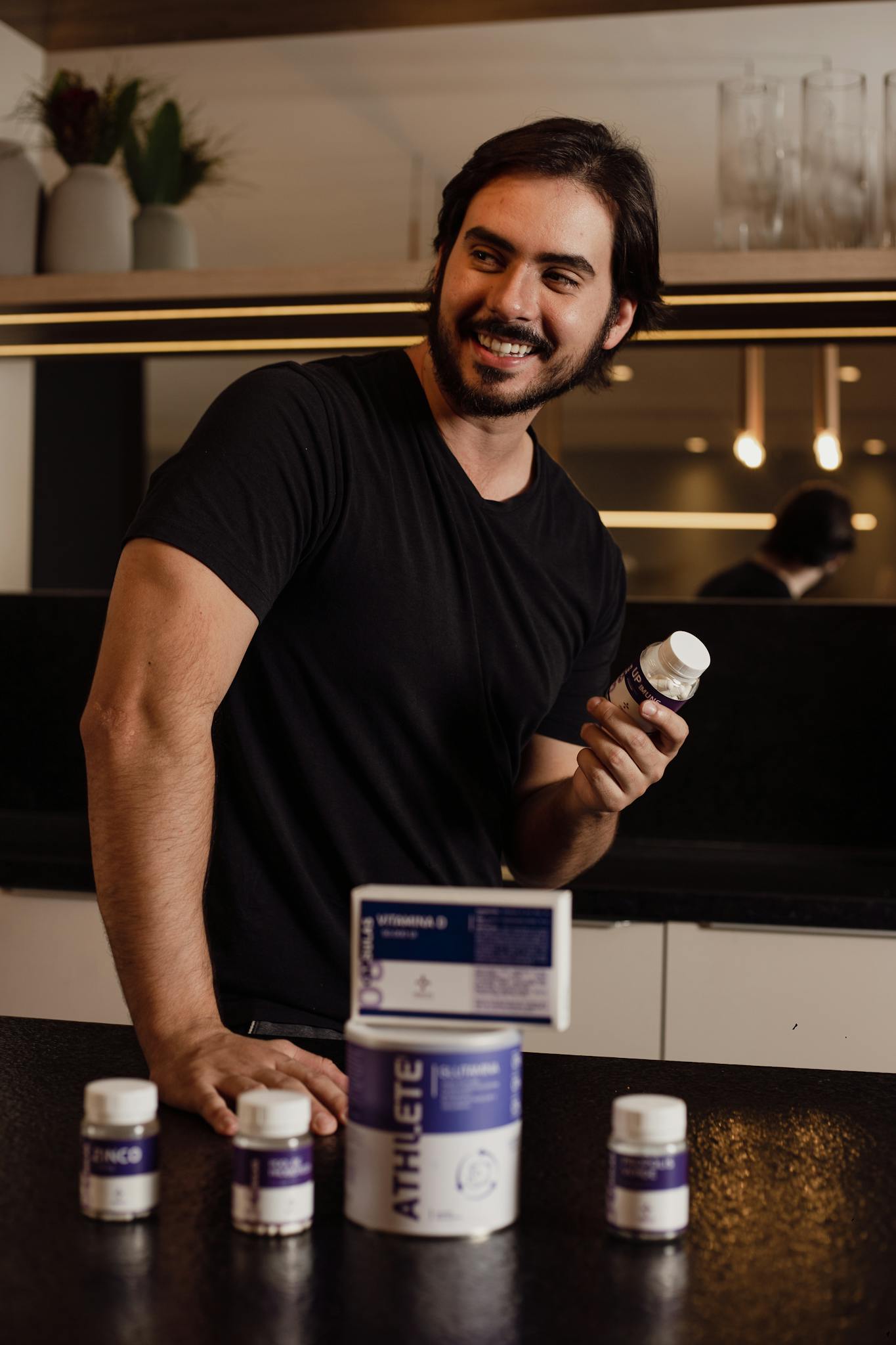 A man in a kitchen holding a supplement bottle and smiling, showcasing vitamin products.
